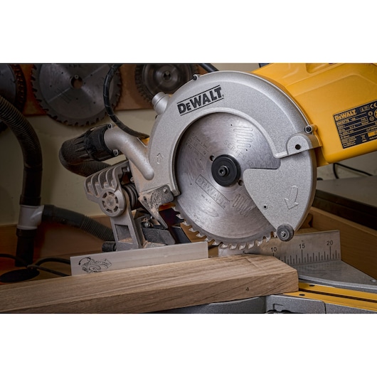 Close-up view of a DEWALT DWS778 miter saw in a workshop, cutting a piece of wood. The saw blade and measurement guide are visible, along with other saw blades mounted on the wall in the background.