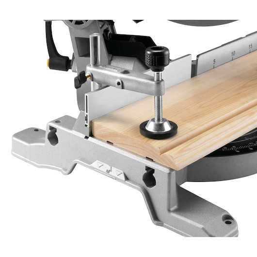 Close-up view of a DEWALT mitre saw with a clamp securing a piece of light-colored wood on the saw base, ready for precision cutting.