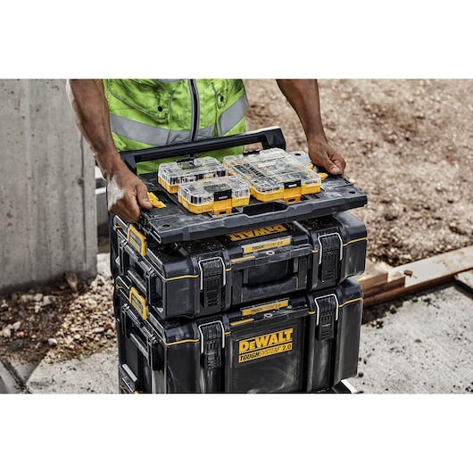 A person in a high-visibility vest stacks several black DEWALT ToughSystem 2.0 toolboxes and organizers outdoors on a worksite.