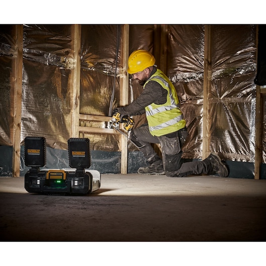 A person wearing a yellow construction helmet and a reflective safety vest is kneeling on a construction site, using a DEWALT power tool. In the foreground, a DEWALT ToughSystem radio is placed on the floor. The background shows wooden framing and insulation materials.
