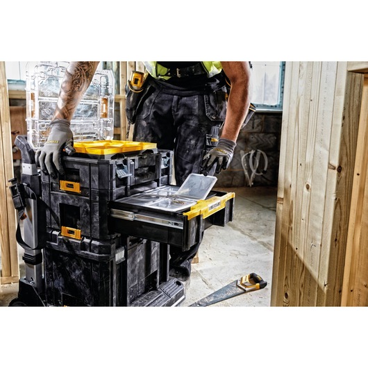 A worker wearing gloves is using a DEWALT stackable tool storage system with multiple compartments and drawers in a construction setting. Various tools and clear storage cases are visible, and the worker is accessing one of the storage drawers.