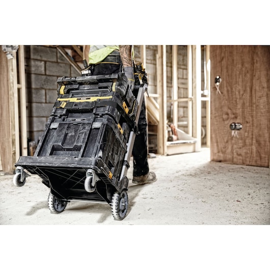 A person in work clothing is using a DEWALT rolling tool box trolley with stacked black and yellow tool boxes on a construction site with unfinished walls.