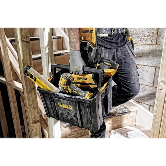 A person wearing work gloves and holding a DEWALT tool box filled with various DEWALT power tools and equipment. The scene takes place at a construction site with wooden framing and concrete walls visible.