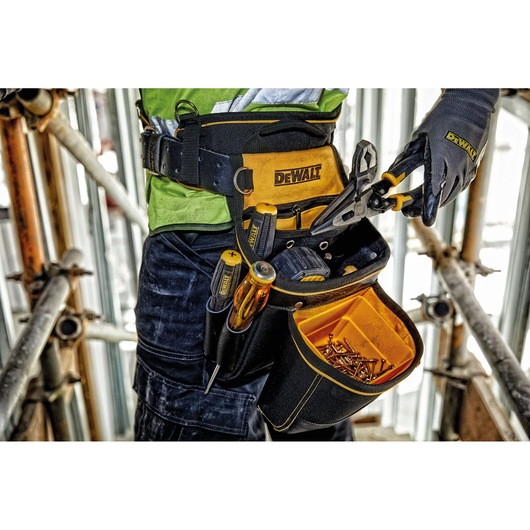 Close-up of a DEWALT tool belt worn by a person on a construction site. The belt holds various tools such as screwdrivers, pliers, and a compartment filled with screws. The person is wearing work gloves and holding a pair of pliers.
