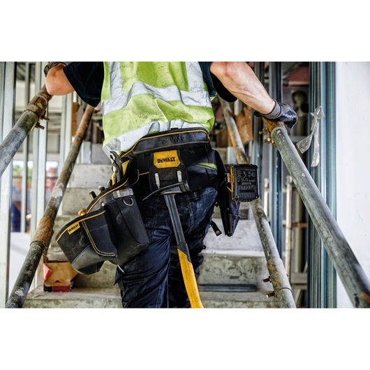 A construction worker wearing a high-visibility vest and a DEWALT tool belt climbs a set of stairs at a worksite. The tool belt holds various tools and equipment useful for the job.