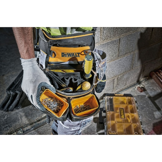 Close-up view of a DEWALT tool belt worn by a person, featuring multiple compartments with tools, screws, and nails. The person is wearing work gloves and standing near a DEWALT storage organizer on a construction site with a brick wall.