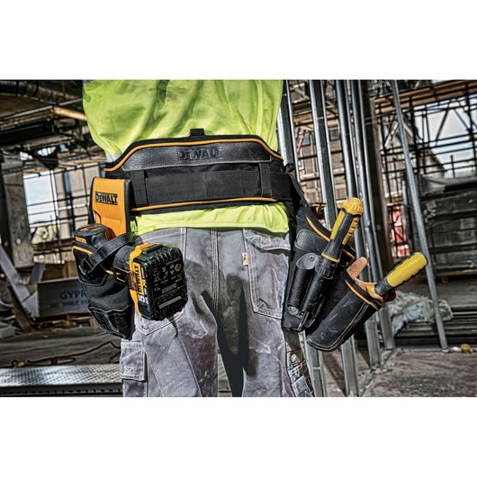 A close-up of the back of a worker wearing a DEWALT tool belt, loaded with various tools and accessories, in a construction site setting.