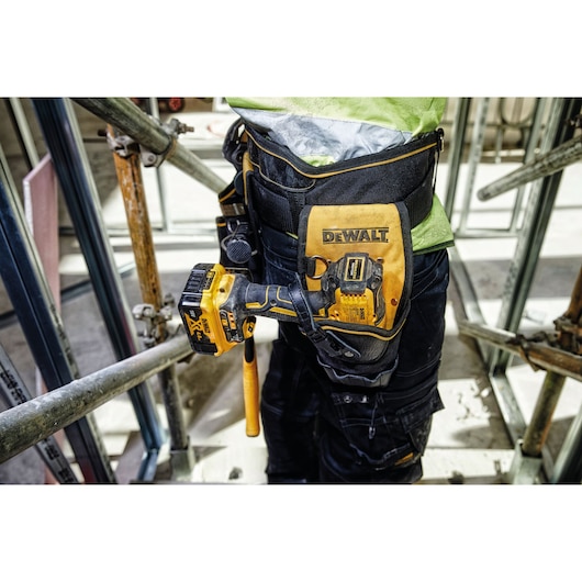 Close-up of a worker wearing a DEWALT tool holster attached to a tool belt. A yellow DEWALT cordless drill is securely placed in the holster. The worker is standing on a construction site with metal scaffolding.