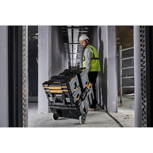 A person wearing a safety vest and hard hat pulls a stack of DEWALT toolboxes on wheels through a construction site hallway.