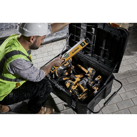 A construction worker wearing a safety vest and helmet is kneeling beside an open DEWALT toolbox filled with various power tools and a spirit level on a paved surface.
