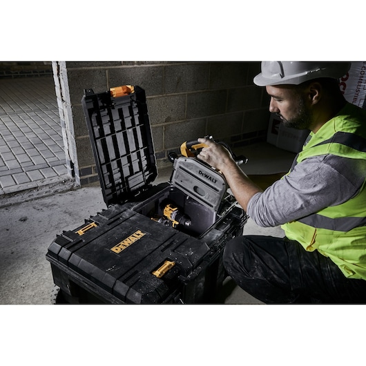 A person wearing a high-visibility vest and a safety helmet is handling a tool inside an open DEWALT storage box, placed on a concrete floor at a construction site. The person's face is intentionally blurred for privacy.