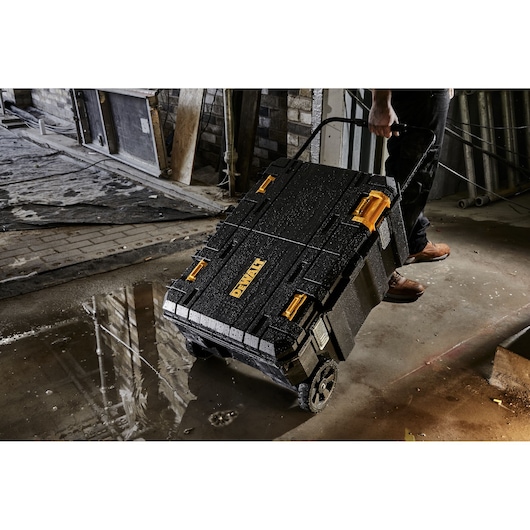A DEWALT rolling tool box with yellow latches, shown in a wet construction site, being pulled by a person.