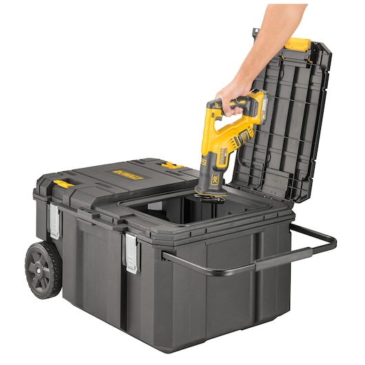 A person is placing a yellow and black DEWALT power tool into a large black wheeled toolbox with an open lid and a pull handle.