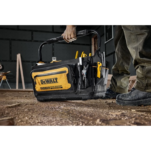 A DEWALT-branded black and yellow tool bag, filled with hand tools, is being carried by a person on a construction site with a dirt floor.
