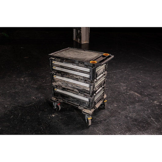 A DEWALT mobile tool storage cart with three stacked drawers, metal slides, and four wheels, placed on a dark floor. The unit is rugged and designed for heavy-duty use in workshops or job sites.
