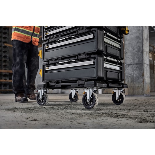 A stack of black DEWALT toolboxes on caster wheels is shown on a rough concrete floor, next to a person wearing work boots, black pants, and an orange safety vest. The environment suggests an industrial or construction site.