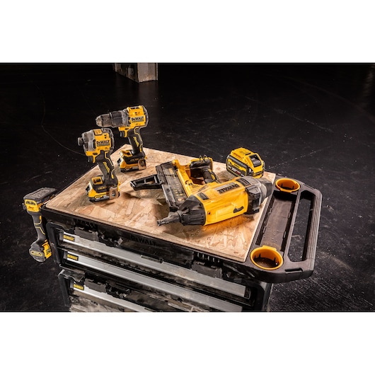 Several DEWALT cordless power tools, including a drill, an impact driver, a nail gun, and a battery pack, are placed on a wooden surface atop a DEWALT tool storage chest in a workshop setting.