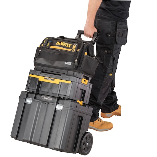 A person wearing work trousers and boots is pulling a stack of DEWALT TSTAK tool storage boxes on wheels. On top is a DEWALT branded tool bag, and underneath are two black and yellow hard tool cases.