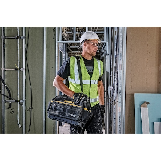 A construction worker wearing a white hard hat, reflective yellow safety vest, and gloves is holding a DEWALT tool bag (DWST82991-1) in a building under construction. The worker's face is deliberately blurred for privacy.