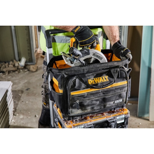 A DEWALT-branded tool bag filled with tools, including a circular saw, is being held by a person wearing work gloves and a high-visibility vest. The tool bag is placed on top of a stack of transparent and black toolboxes on a construction site.