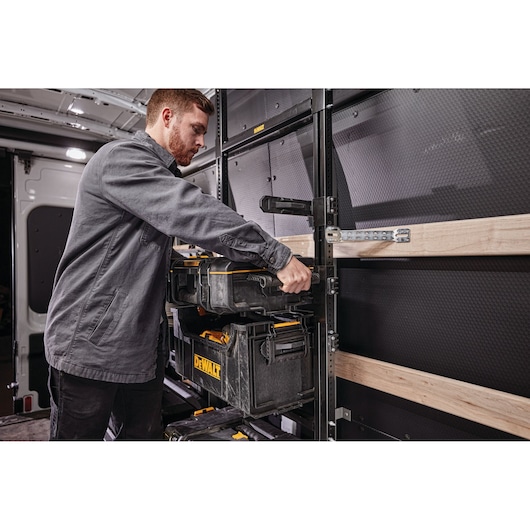 Person placing a DEWALT tool storage box onto a metal shelving system inside a van. The shelving is mounted with wooden slats and other DEWALT toolboxes are visible.