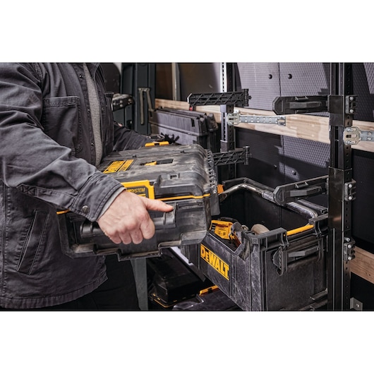 A person is placing a DEWALT ToughSystem tool case onto a rack or shelving unit inside a vehicle. The rack holds multiple DEWALT storage boxes, and tools are visible inside one of the open cases.