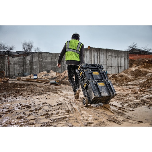 A person wearing a green safety vest and helmet pulls a DEWALT wheeled tool storage box across a muddy construction site, with concrete walls and building materials visible in the background.