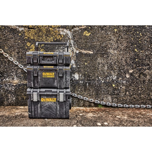 Stack of three DEWALT ToughSystem 2.0 tool storage boxes placed outdoors against a rough textured wall, with each box featuring the yellow DEWALT logo and heavy-duty latches.