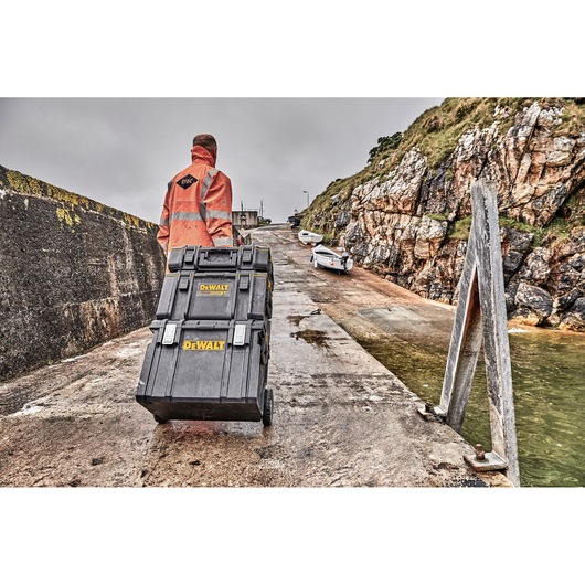 A person wearing a high-visibility jacket is pulling a stack of two DEWALT black toolboxes with yellow logos on wheels along a sloped concrete path near water, with rocky cliffs and boats in the background.