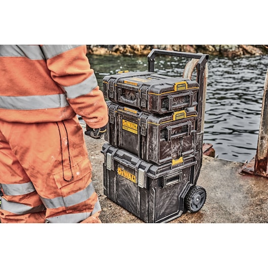 A stack of three DEWALT toolboxes on wheels is shown next to a person wearing an orange high-visibility work suit, positioned on a dock near a body of water.