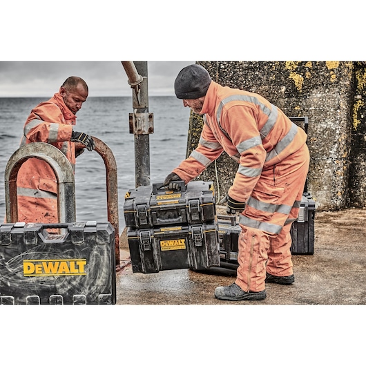 Two individuals wearing orange high-visibility work suits are handling DEWALT ToughSystem toolboxes outdoors near a body of water. The toolboxes are black with yellow DEWALT branding, and the scene appears to take place on a rough, industrial surface.