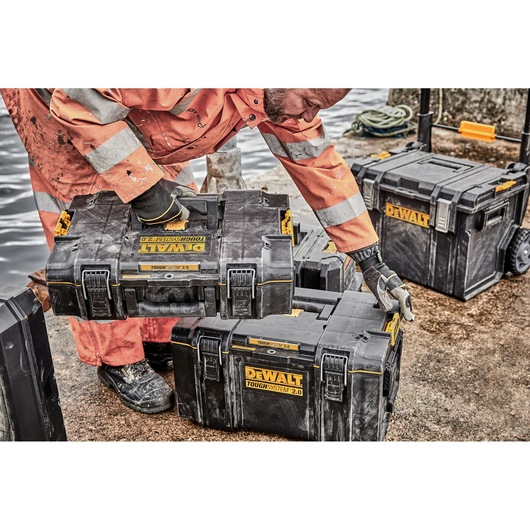 A person wearing an orange high-visibility suit and gloves is handling a DEWALT ToughSystem 2.0 toolbox on a construction site. Several DEWALT toolboxes are visible, with one stacked on top of another and one larger toolbox on wheels in the background.