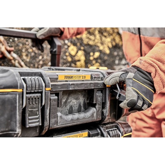 Close-up of a person wearing gloves and an orange jacket, handling a DEWALT ToughSystem 2.0 tool storage box outdoors.