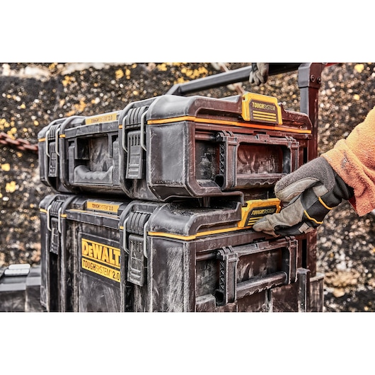 Two DEWALT ToughSystem 2.0 toolboxes stacked outdoors, with a person wearing work gloves and an orange jacket interacting with the latch on the lower box.
