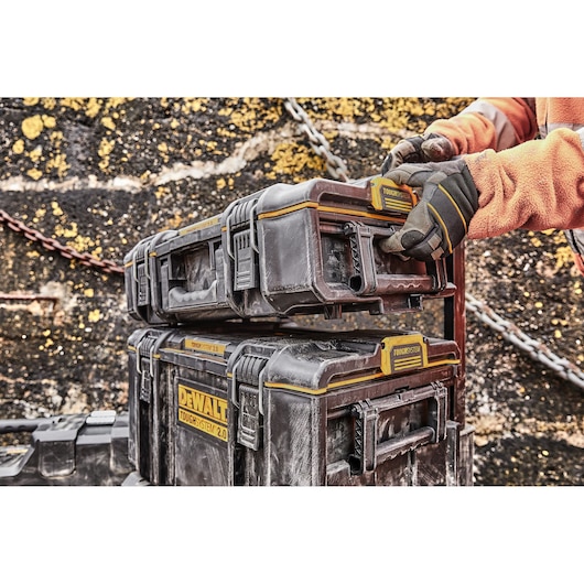A close-up of a person wearing gloves organizing stacked DEWALT ToughSystem 2.0 storage boxes outdoors, with a weathered wall and chain in the background.