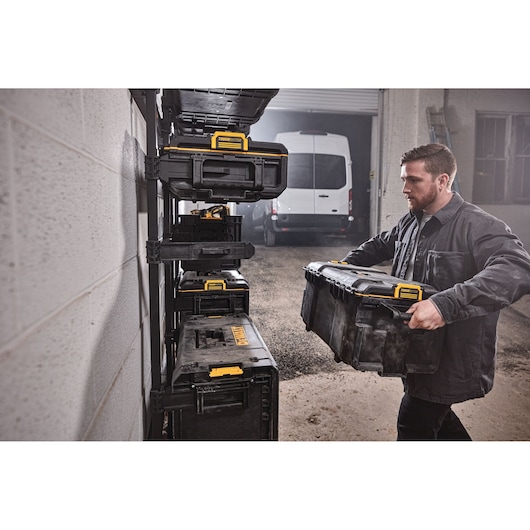 A person is placing a DEWALT ToughSystem storage box onto a wall-mounted rack with several other similar yellow and black DEWALT toolboxes. The scene is in a garage or workshop, and a white van is visible in the background.