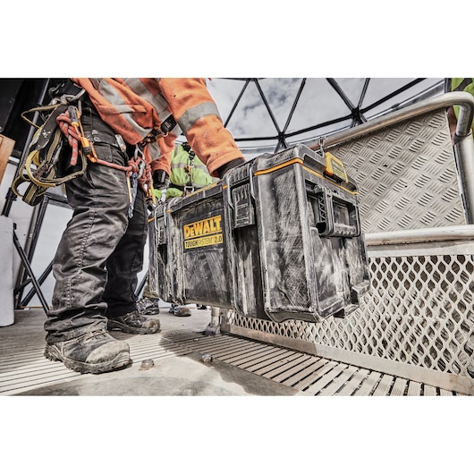 A worker wearing safety gear is carrying a dirty DEWALT ToughSystem 2.0 tool box up a metal staircase at a construction site. The focus is on the tool box and the worker's lower body, highlighting rugged outdoor use.