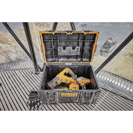 A DEWALT ToughSystem 2.0 storage case with the lid open, containing two yellow and black DEWALT power tools and a pair of gloves, placed on a metal platform near a cliff and body of water.