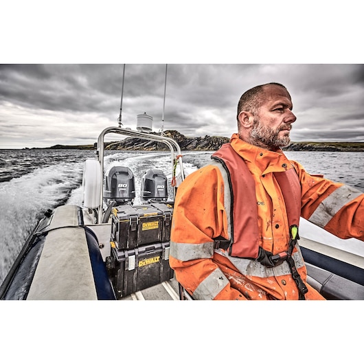 A DEWALT DWST83294-1 tool storage box is shown secured on a boat. In the foreground, a person wearing an orange high-visibility protective jacket and life vest is seated at the helm. The boat is moving on water with land visible in the background under a cloudy sky.