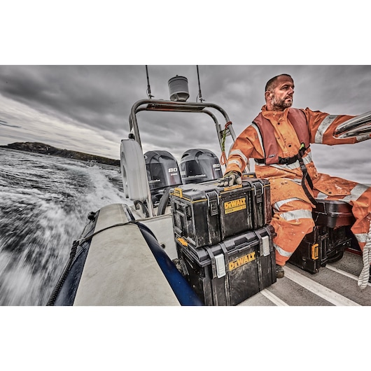 A person dressed in orange waterproof gear is sitting on a moving motorboat. Two black DEWALT toolboxes are stacked next to the person, placed on the floor of the boat. The boat is traveling at high speed, creating water spray behind it, under a cloudy sky.
