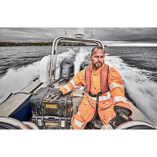 A person wearing an orange safety suit and life jacket is sitting on a speeding boat, holding two DEWALT toolboxes. The boat is moving quickly across the water, with waves visible behind it and a cloudy sky overhead.