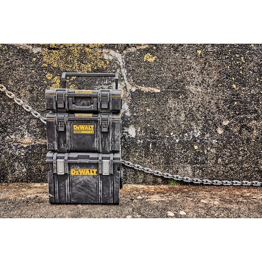 Three stacked DEWALT ToughSystem 2.0 tool storage boxes are positioned outdoors against a rough concrete wall with a chain running behind them. The boxes are black with yellow DEWALT branding.