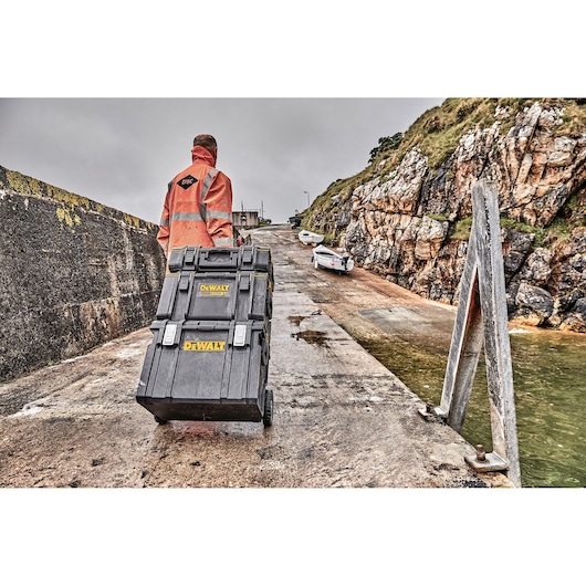 A person in an orange safety jacket is rolling a stack of DEWALT toolboxes on wheels along a wet concrete dock near boats and rocky cliffs.