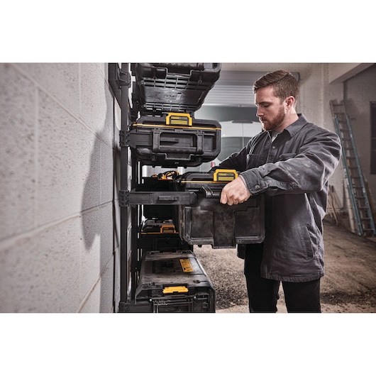 A person with a blurred face organizes DEWALT black and yellow toolboxes on a wall-mounted storage rack in a workshop setting.