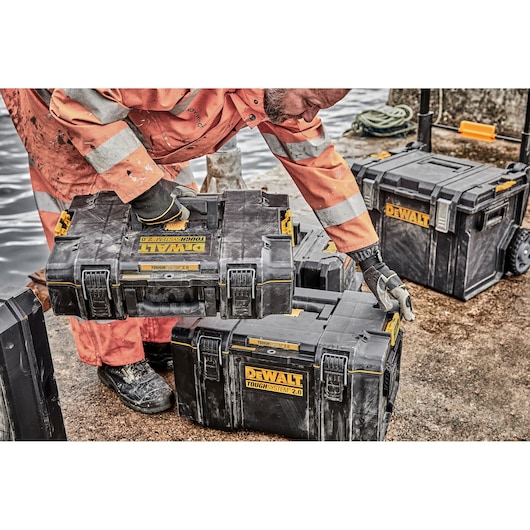 A worker in an orange safety suit handling DEWALT ToughSystem 2.0 storage boxes outdoors, with several black and yellow toolboxes on the ground and a larger box on wheels in the background.