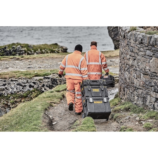 Two individuals wearing orange high-visibility work suits are outdoors near a stone wall, pulling DEWALT stacking tool storage boxes on wheels along a grassy and rocky path.