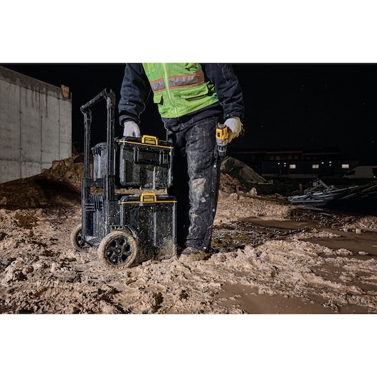 A construction worker wearing a green safety vest stands on a muddy worksite at night, holding a portable power tool and beside a stack of DEWALT storage boxes mounted on a wheeled trolley.