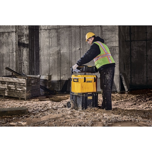 A person wearing a hard hat and safety vest is handling a stack of DEWALT toolboxes with wheels in a muddy construction site.