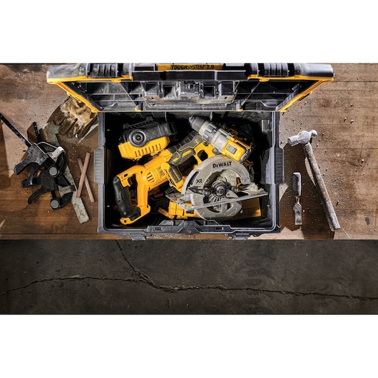 An open DEWALT ToughSystem 2.0 storage box containing several yellow and black power tools, including a circular saw and a drill, on a workbench with hand tools and clamps nearby.
