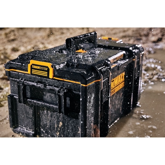 A DEWALT ToughSystem tool box, DWST83294-1, is shown outdoors on wet muddy ground, with water splashing across its rugged and waterproof exterior.
